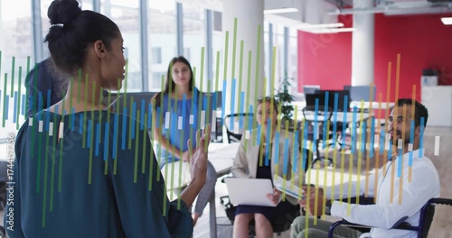 Presenting woman in green blouse gesturing at charts in office, with laptop wheelchair, copy space