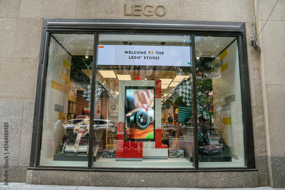 Naklejka premium New York City, New York, USA - 07.31.2024: A wide-angle view of a LEGO store window shows a racing-themed setup with checkered flags and a large digital Welcome to the LEGO Store sign. The glass