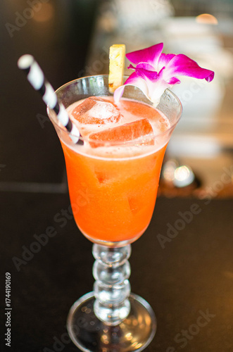 A colorful tropical cocktail served in a tall glass with ice, striped straw, purple orchid flower, and pineapple slice on a dark background.