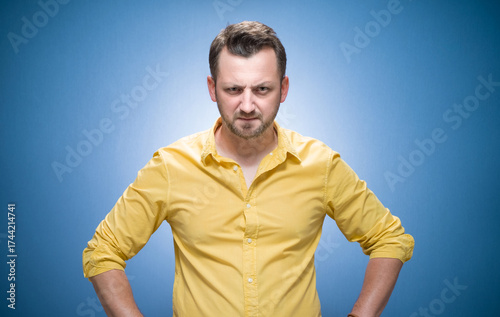 Bossy man staring to you over blue background, dresses in yellow shirt. Jealous guy looking with frowning expression