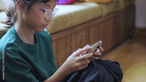 Asian kid, a young girl, is playing a game on her mobile phone at home. This shows child interacting with smartphone technology, highlighting focused entertainment and modern childhood.
