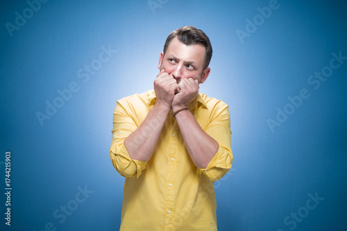 Worried young man biting her fingernail, she know the secret over blue background, dresses in yellow shirt