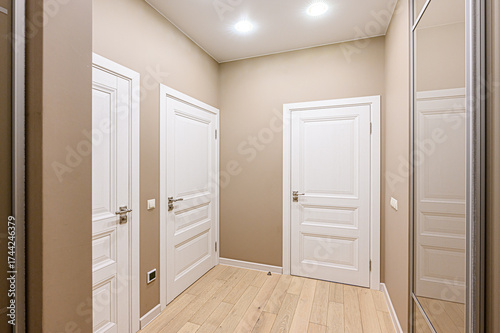 A well-lit hallway with beige walls, light wood flooring, and white doors. There are two ceiling lights and a mirror closet on the right side of the wall