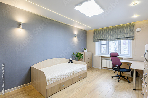 A cozy room features a bed, desk, and natural light. The room has a grey wall, wooden floors, and modern lighting