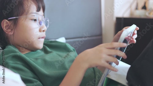 Selective focus of Asian kid girl, playing game on tablet with concentration. Concepts of children technology use, online learning, screen time, digital lifestyle, and modern childhood education.