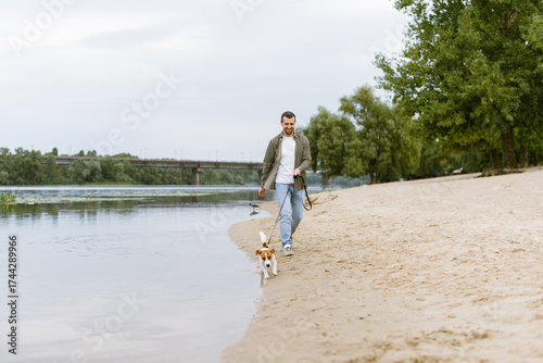 Smiling man walking with Jack Russell Terrier dog on sandy beach near river in the morning