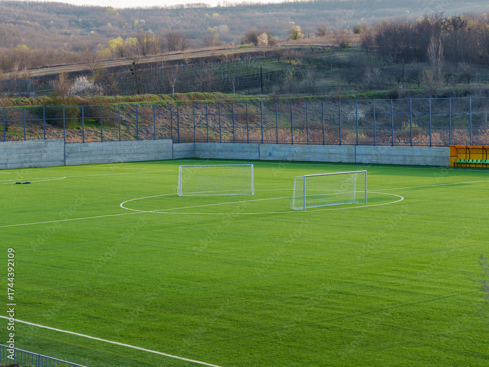 Naklejka premium A soccer field sits empty under a clear sky with two goals at either end. The bright green grass contrasts with the surrounding hills, showcasing a peaceful afternoon scene.