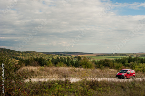 Dirt road with car into a landscape with hills of dry grass, trees, forests o...