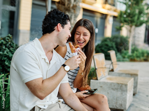 Happy couple  eating a pastry croissant, smiling and looking away on a city street