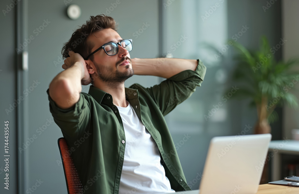 Fototapeta premium Young man with glasses sits at desk with laptop, hands behind head eyes closed, taking a break. Person relaxes in office workplace, feeling calm.