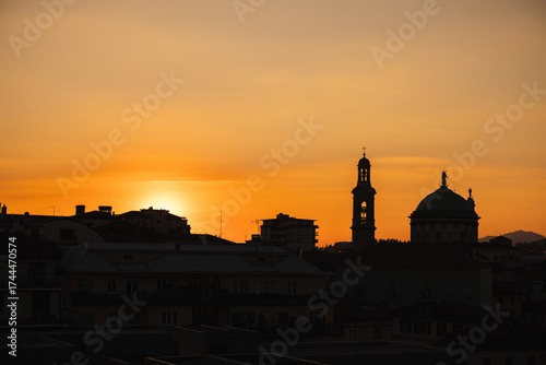 Cityscape of Bergamo, Italy at sunset with silhouettes of historical buildings and church domes against a golden orange sky