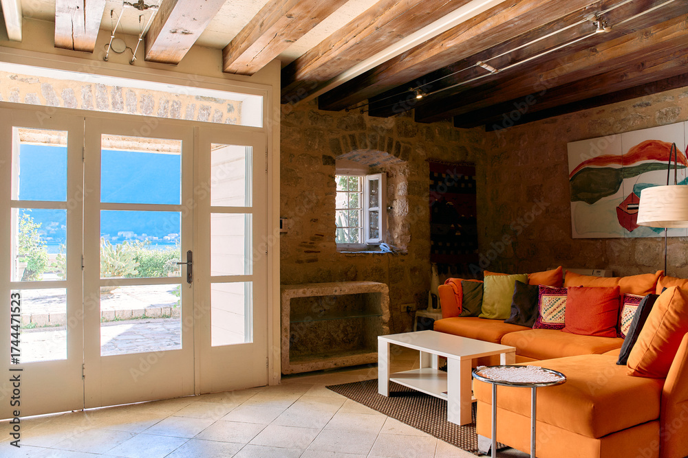 Obraz premium Cozy Living Room with Bright Orange Sofa Overlooking Scenic View Through Open Doors in a Stone-Finished Home