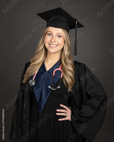 portrait of a young blonde female graduate wearing cap and gown isolated on grey background