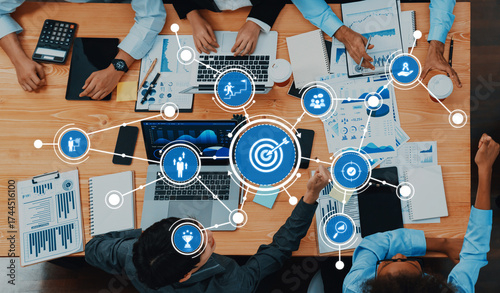 Photos A diverse group of professionals engages in a strategy meeting, analyzing data on laptops and charts, focusing on targets and project outcomes in a modern workspace