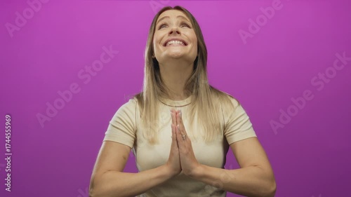 Woman smiling hopefully with hands in praying gesture against vibrant pink background under isolated light setting.