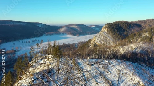 An aerial shot of a winter landscape in the South Ural Mountains, Russia. The footage shows snow capped hills and a frozen river in a valley, with a clear blue sky above. The foreground features a slo