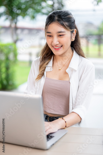 Free Download Smiling young adult asian woman looking and using a laptop while sitting at wooden table in the park - GFXLoad