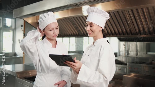 Two White female gourmet chefs in aprons discuss ingredient preparation and menu on tablet, cooking in kitchen, professional partners in delicious cuisine,  in catering restaurant, and culinary meals.