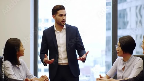 Young leader standing at meeting room with his team around conference table of experts and having speech