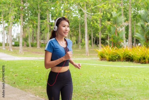 Smiling young asian woman enjoying refreshing jog in lush green park listening to music with her headphone. Perfect for health & wellness.