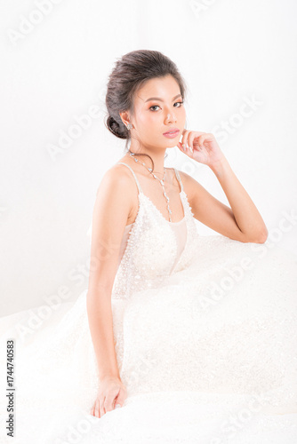 Beautiful Asian woman in a white wedding gown and elegant jewelry poses gracefully against a clean studio shot on white background, exuding timeless beauty.