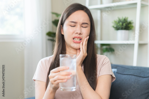 Suffering from toothache, hurt asian young woman touch cheek, face expression ache or feel pain, sensitive molar teeth, hand holding glass of water with ice, inflammation when drink cold, healthcare.