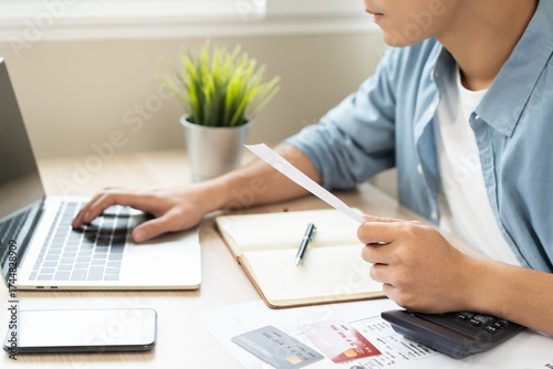 Stressed asian young businessman, employee male using laptop computer to calculate expenses, hand holding bills and receipt for to payment on table at home. Financial, finance of banking concept.