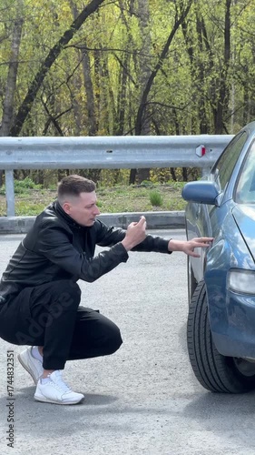 Caucasian man looks at and photographs deformation on his car body. 