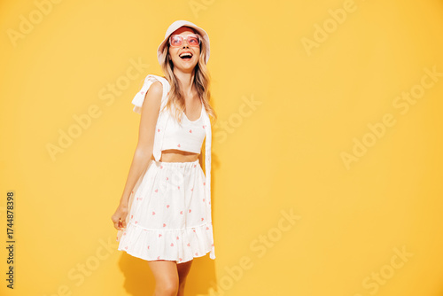 Young beautiful smiling blond female in trendy summer  white top and skirt clothes. Carefree woman posing near yellow wall in studio. Positive model in panama hat. Cheerful and happy