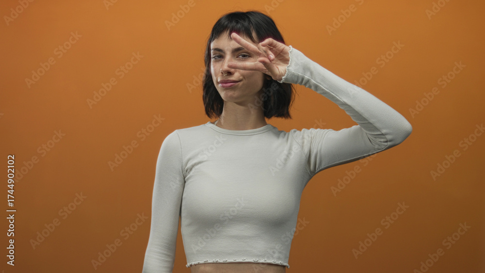 Fototapeta premium Woman wearing a white crop top in a casual pose smiling and making a peace sign with her hand by her eye in orange studio; joy.