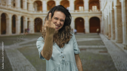 Woman showing middle finger in a sunlit building courtyard with arched corridors and worn stone floors; defiance.