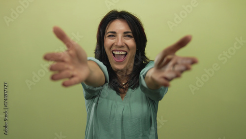Hispanic woman with broad smile extends bare arms forward for hug gesture in green studio; affection.
