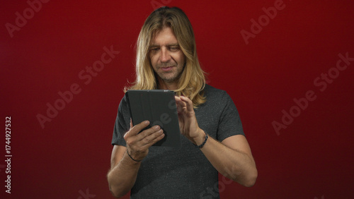 Man with long blond hair tapping tablet touchscreen with finger in a red studio; digital engagement focus.
