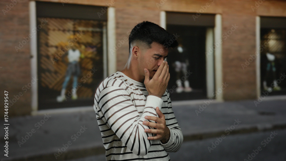 Fototapeta premium Young man with striped shirt standing on city street looking anxious in urban outdoor setting with hands near face expressing emotions under brick building backdrop.