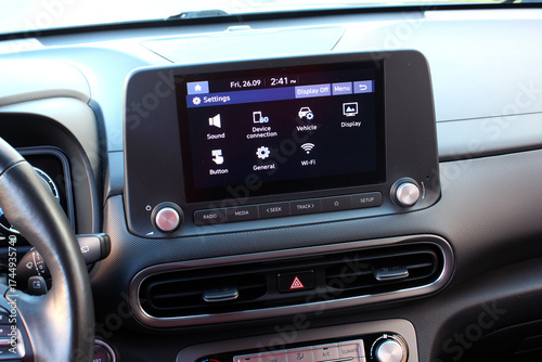 Car infotainment touchscreen displaying settings menu with icons for sound, Wi-Fi, and vehicle options. Dashboard features matte black finish with silver knobs and control buttons. Car multimedia.