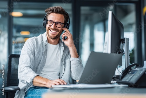 A man with headphones is at a desk using a laptop and phone