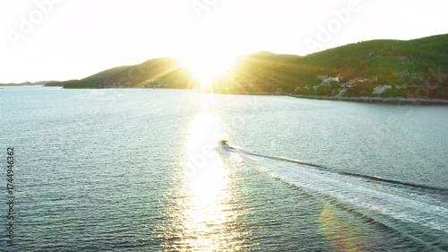 A motorboat cruises along a scenic coastline, crossing the sun's reflection on the water at sunset. This aerial shot embodies a luxury lifestyle, summer vacation, and high-speed water recreation.