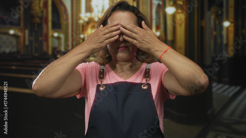 Middle age hispanic woman in pink shirt and apron covers eyes with hands in church building; serenity.