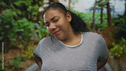 Woman wearing striped tshirt holding lower back on park walkway surrounded by lush greenery and tall trees, frowning with furrowed brow; pain.