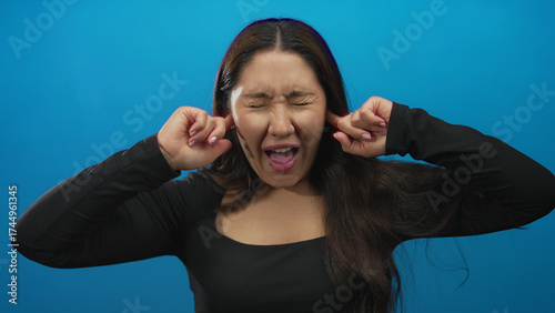 Young hispanic woman with long dark hair covers ears with hands on a vibrant blue studio set; annoyance.