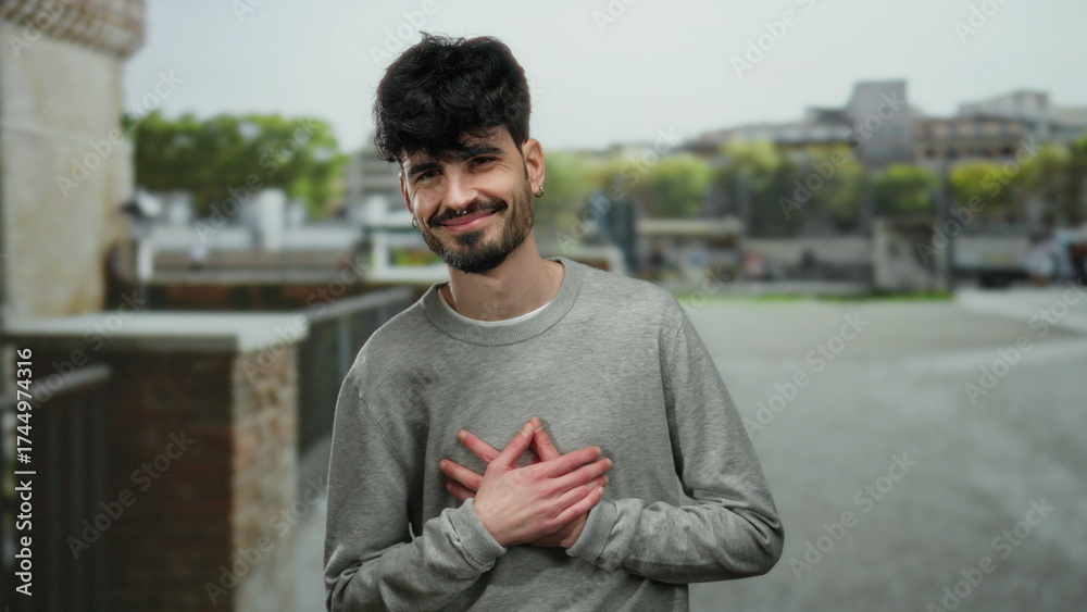 Fototapeta premium Young man with hands on chest smiling warmly outdoors in urban street setting.