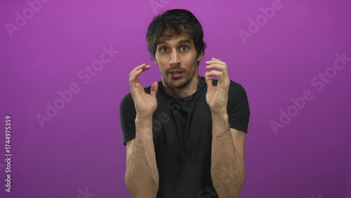Young man raising hands defensively in studio with purple wall and tense expression and hunched posture; anxiety.