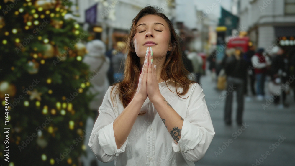 Fototapeta premium Woman praying outdoors on city street near christmas tree surrounded by people in festive setting wearing white shirt in urban area during holiday season.