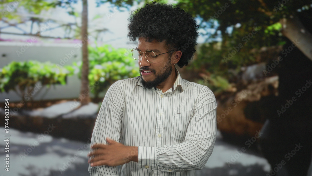 Fototapeta premium Man wearing glasses and striped shirt stands outdoors with trees in the background, expressing thoughtful mood on a sunny day.