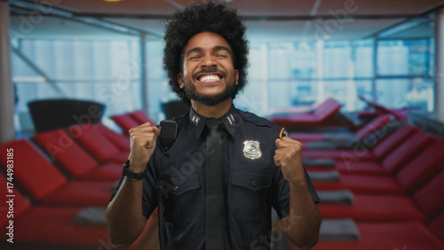 Smiling african american police officer in uniform celebrates indoors on a cruise boat with vibrant red seating, capturing joy and professionalism at sea.