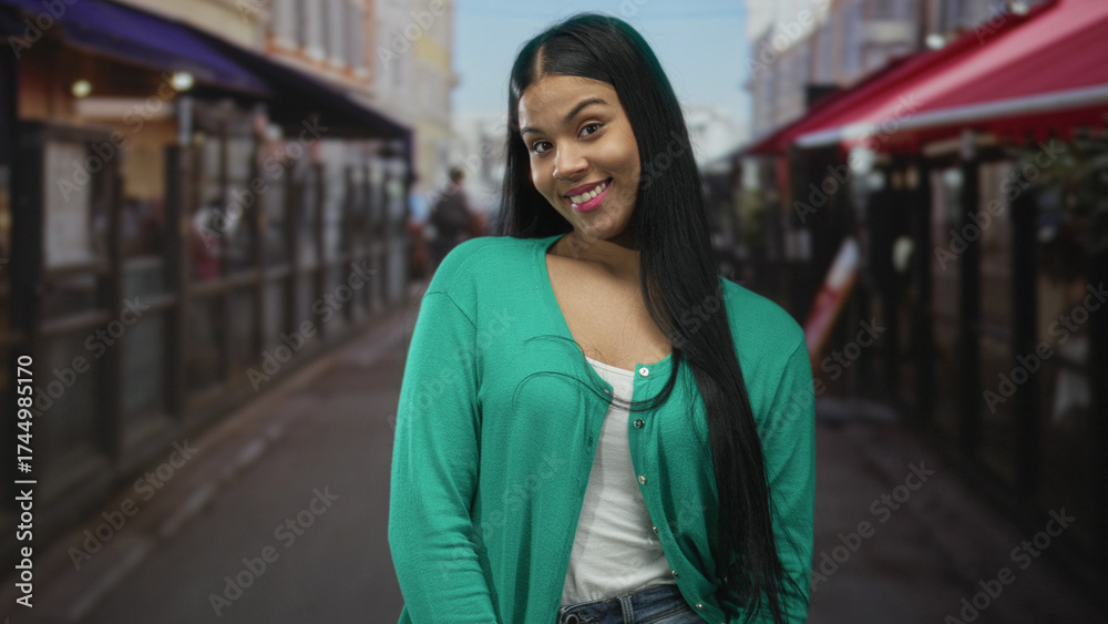 Fototapeta premium Latin woman with long dark hair in green cardigan smiles on city street outside cafe; confidence joy.