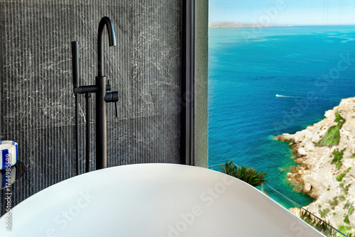 Luxury bathroom with freestanding bathtub and ocean view. Dark stone tiles, black faucet, and modern minimal style create relaxing spa atmosphere above turquoise coast.