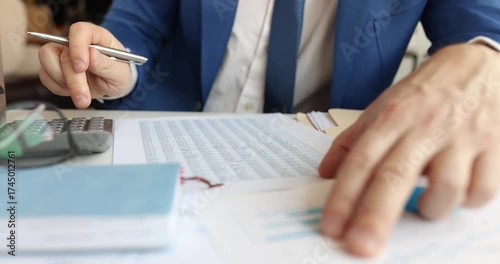 Man in suit studies printed accounting report on desk with pen. Analyst marks figures highlighting rows and checking totals for update sheet