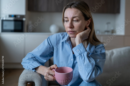 Sad middle aged woman sits in living room deep in thoughts. Melancholy European female feeling tired, worried, drinking tea in reflective mood. Loneliness, emotionally drained, pondering life issues