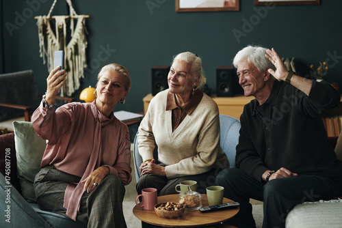 Three Caucasian seniors sitting together, smiling and posing for selfie with smartphone, older man waving hand while two senior women looking at camera, drinks and snacks on table